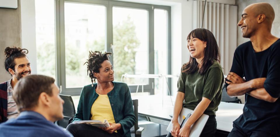 A group of colleagues gathered in a conference room or work area talking and smiling. 