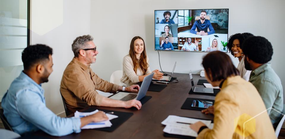 A group of coworkers are holding a meeting in a conference room while several of their colleagues are attending it virtually through a large computer screen.