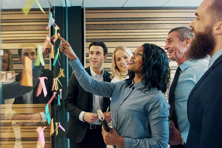 Group of individuals in business setting in front of a board and reviewing sticky notes or a process map.